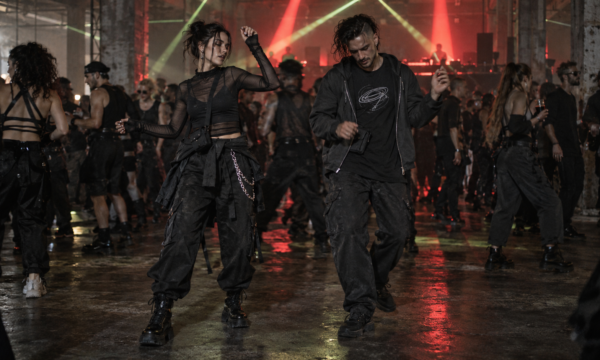 Twee ravers in zwarte outfits en cargobroeken dansen in een industriële hal onder rode en groene lichtstralen.